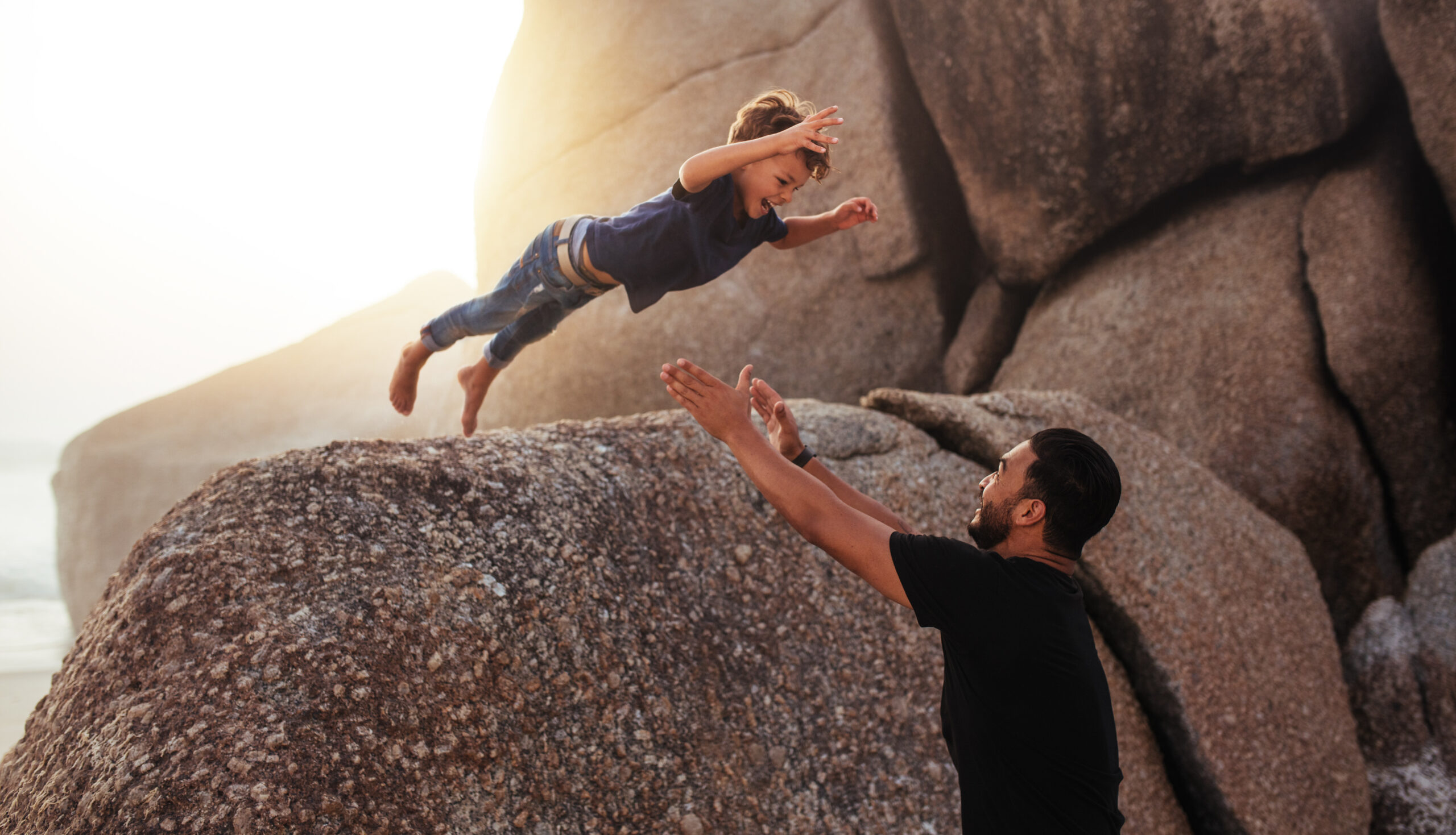 Shot of cute little boy leaping into his fathers arms from a big rock. Father and son enjoying summer holidays at the beach.