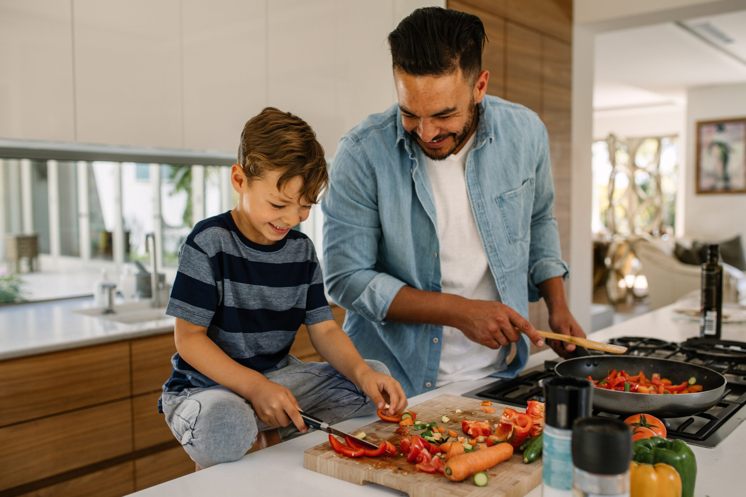 Little boy cutting vegetables while his father cooking food in kitchen. Father and son preparing food at home kitchen.