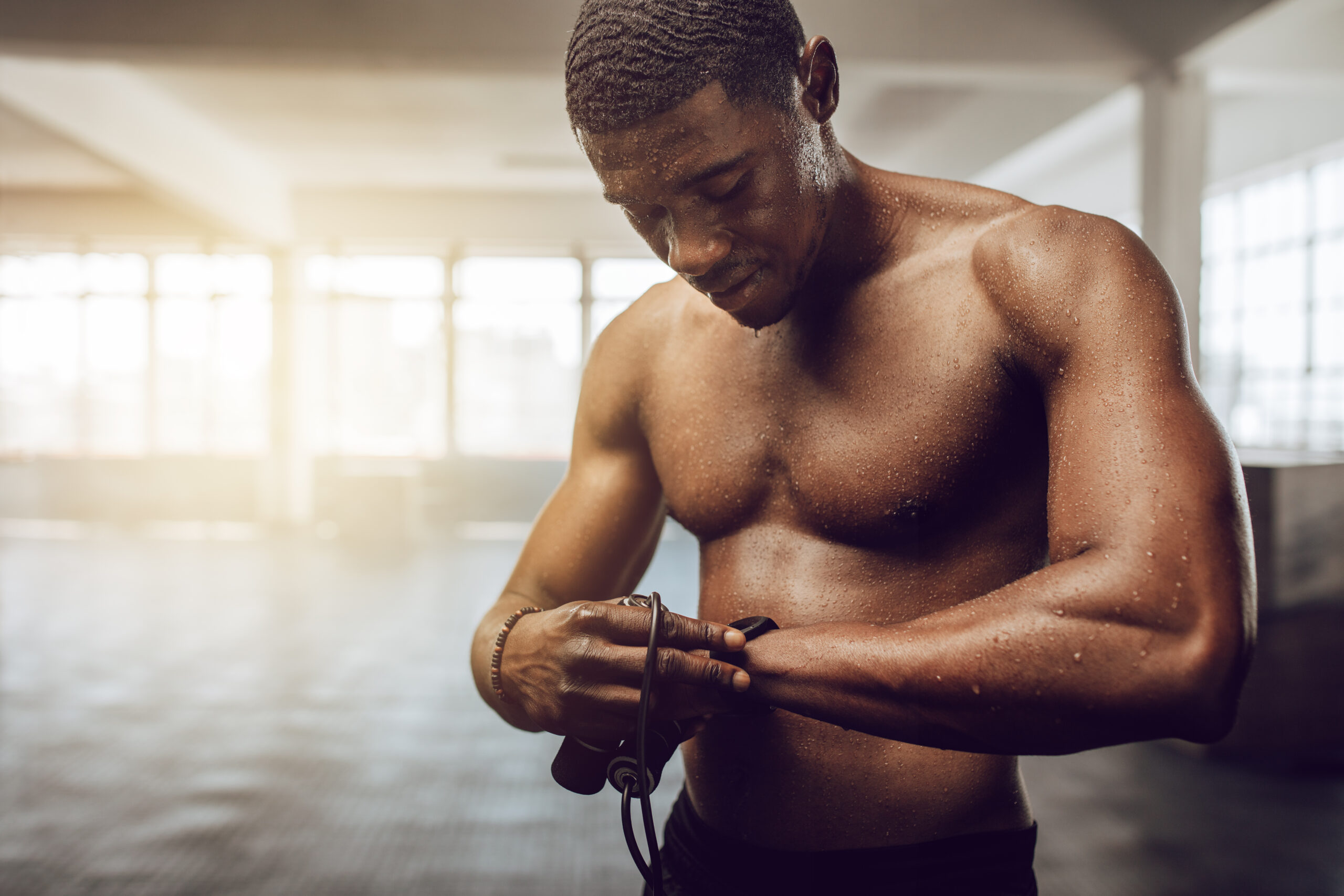 Man setting a timer in his wrist watch standing in the gym. Bare chested muscle athlete holding skipping rope with sweat all over his body after workout.