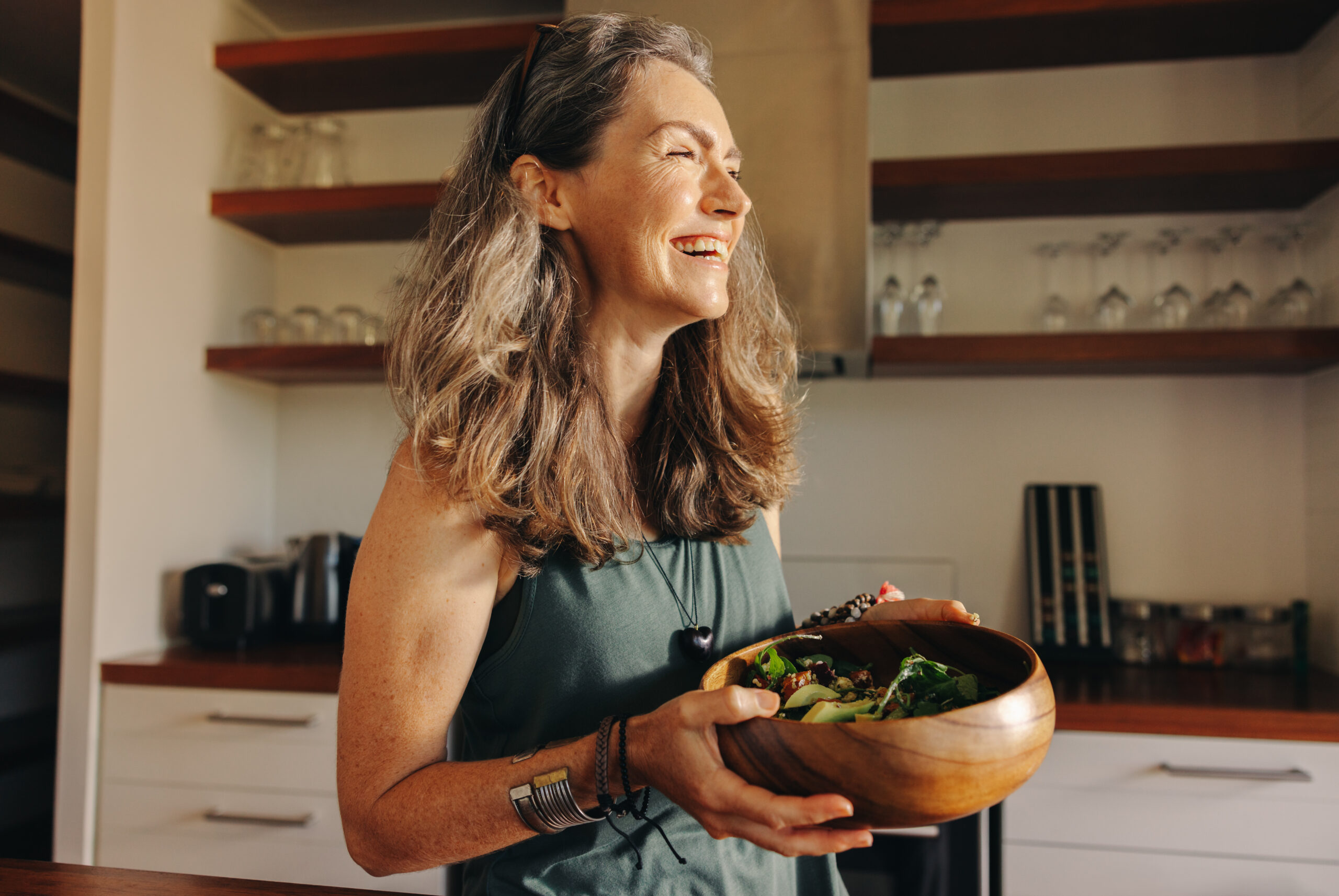 Aging woman smiling happily while holding a buddha bowl in her kitchen. Happy senior woman serving herself a healthy vegan meal at home. Mature woman taking care of her body with a plant-based diet.